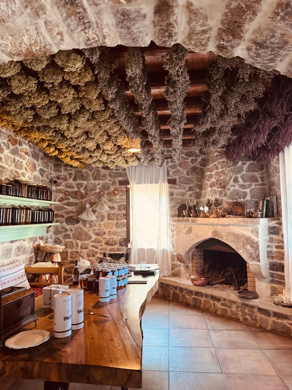 Bundles of dried herbs hanging across a stone room above jars, a wooden table and a fireplace