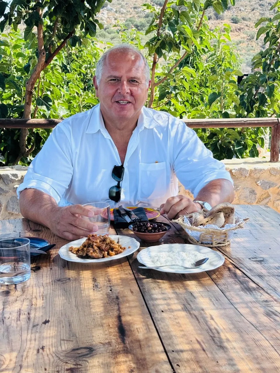 A man in a white shirt seated at a wooden table with cheese, olives and bread