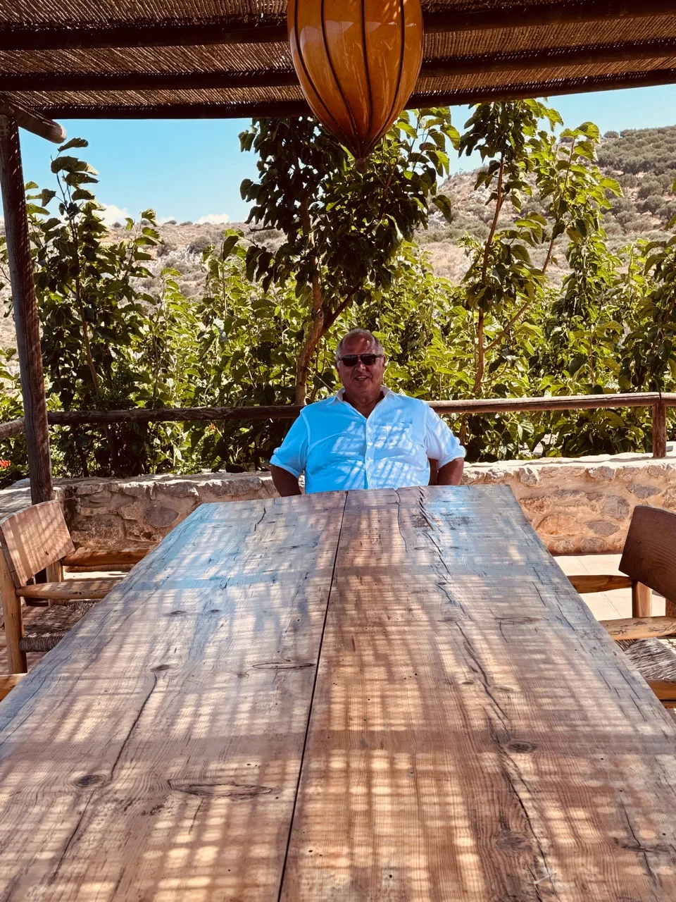 A man wearing sunglasses at the far end of a long wooden table beneath a woven canopy