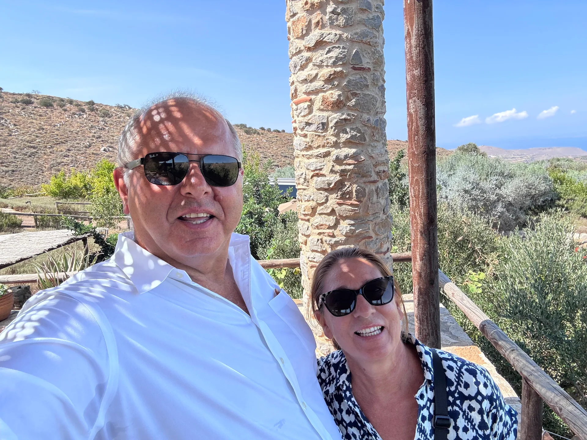 Two visitors in sunglasses taking a selfie beside a stone pillar with olive trees and hills behind