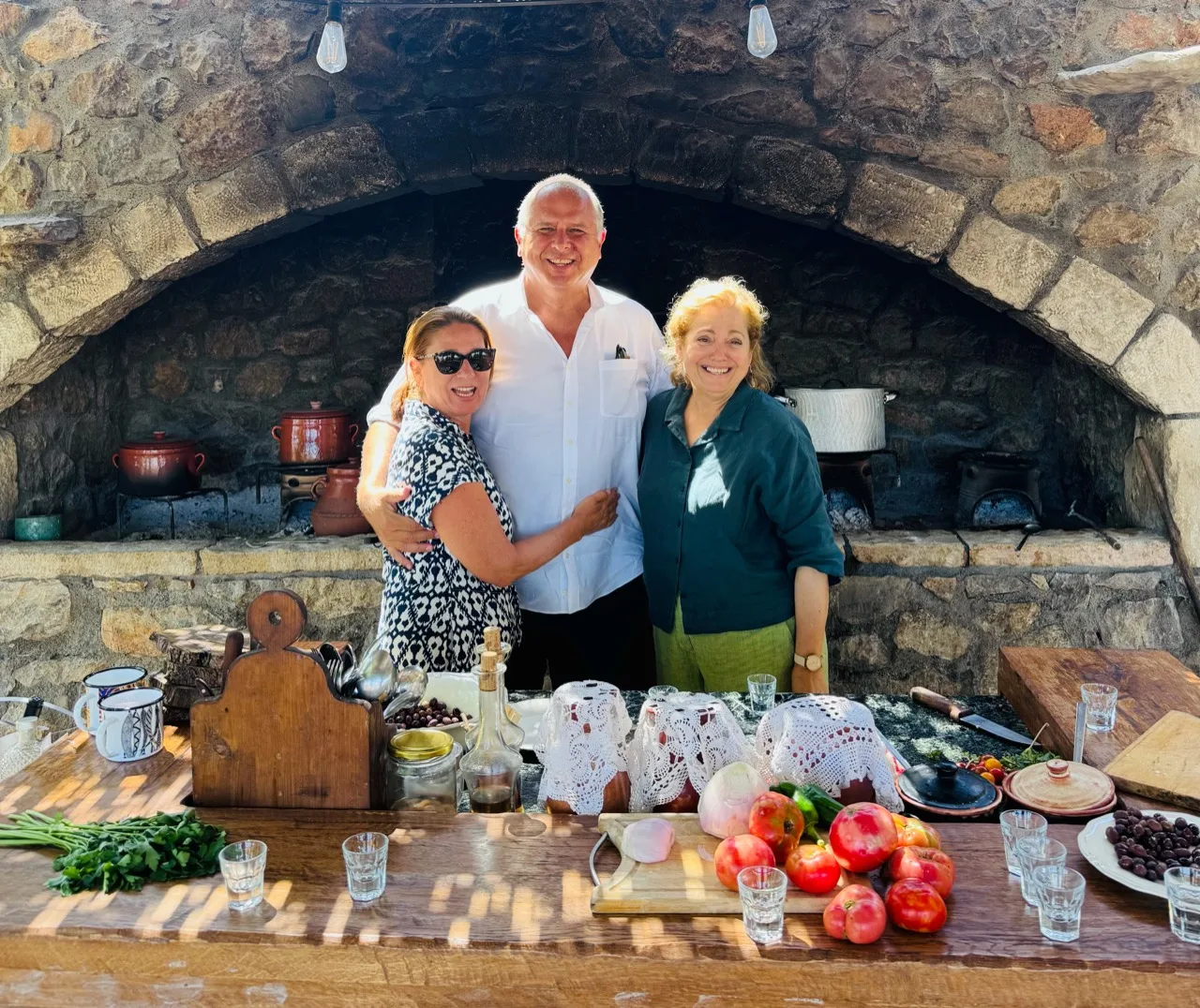 Three people smiling behind an outdoor kitchen counter beneath a large stone cooking hearth