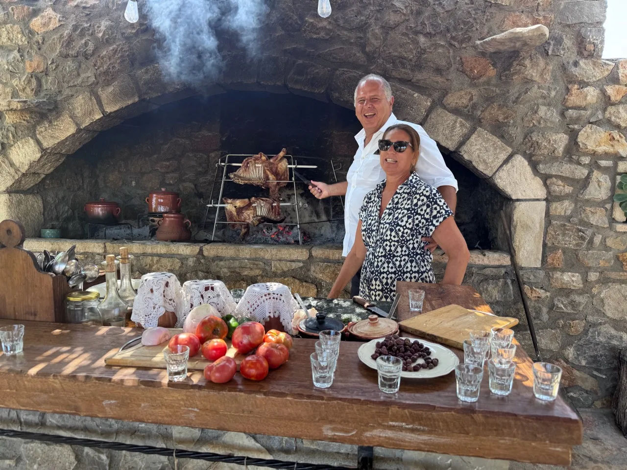 Two visitors smiling beside meat roasting at a stone hearth, with tomatoes and olives on the counter