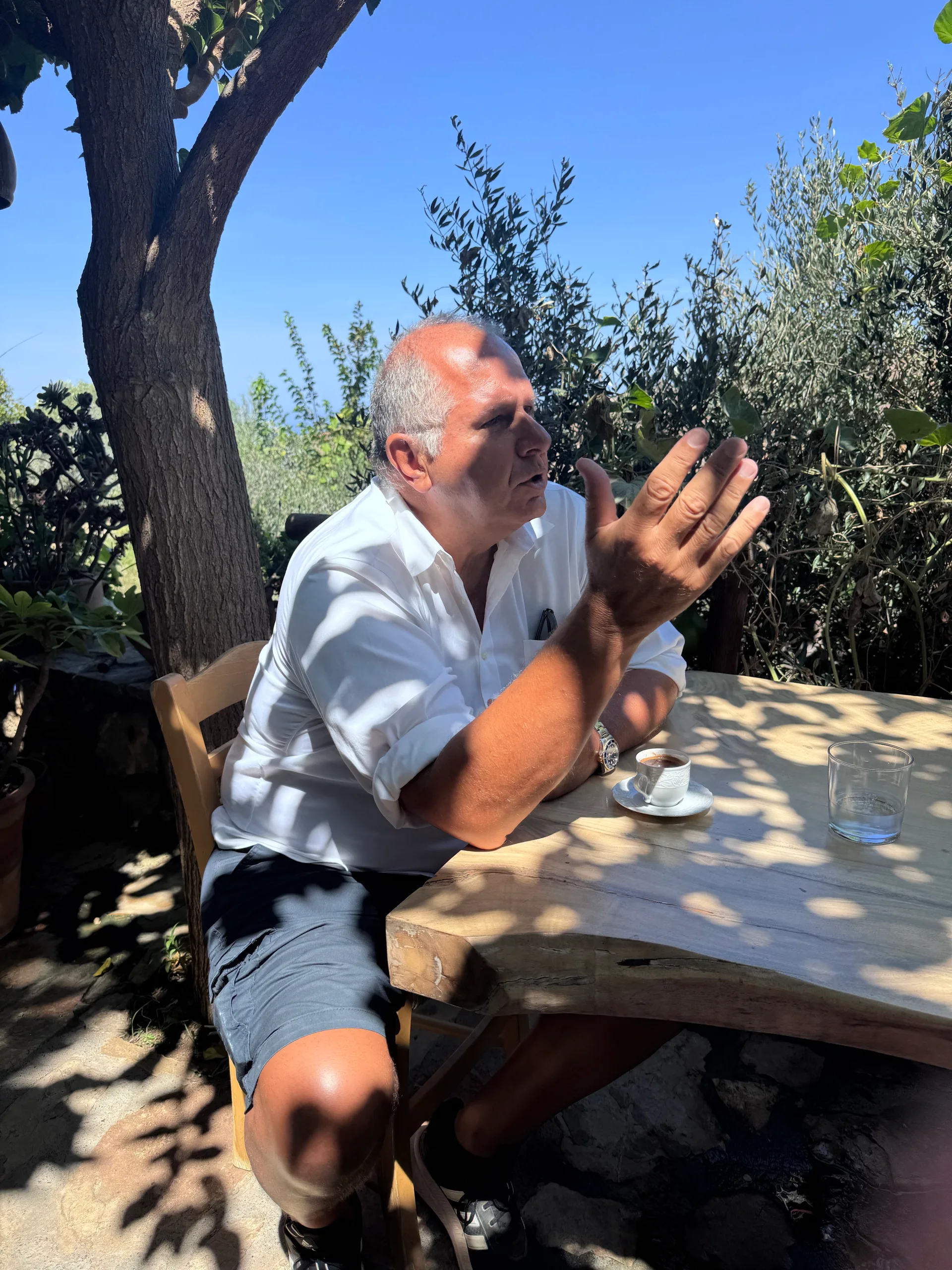 A man in a white shirt gesturing at a shaded outdoor table beside a small coffee cup
