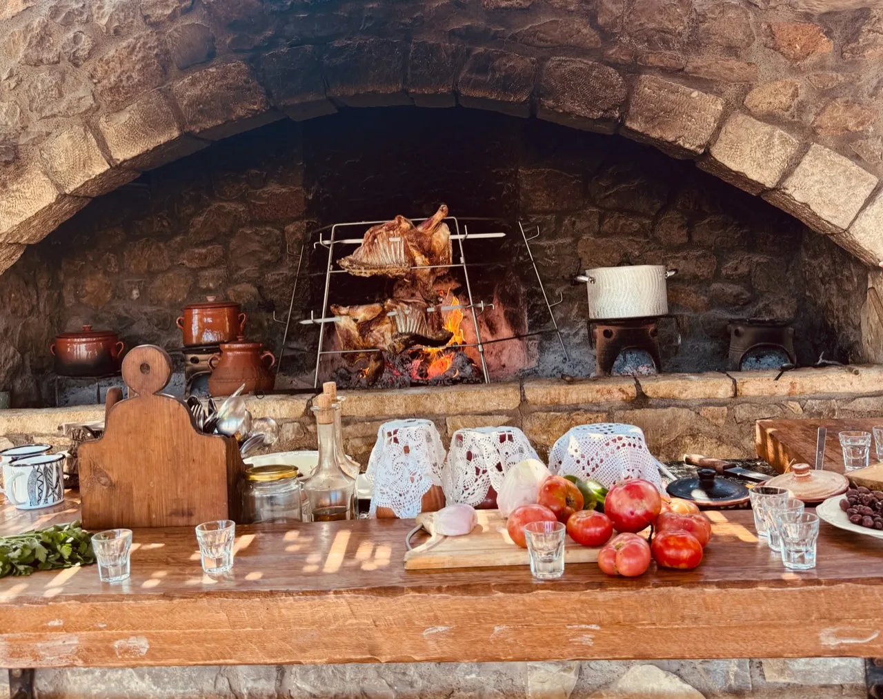 Meat roasting over a fire inside a stone hearth behind a counter of tomatoes, jars and glasses