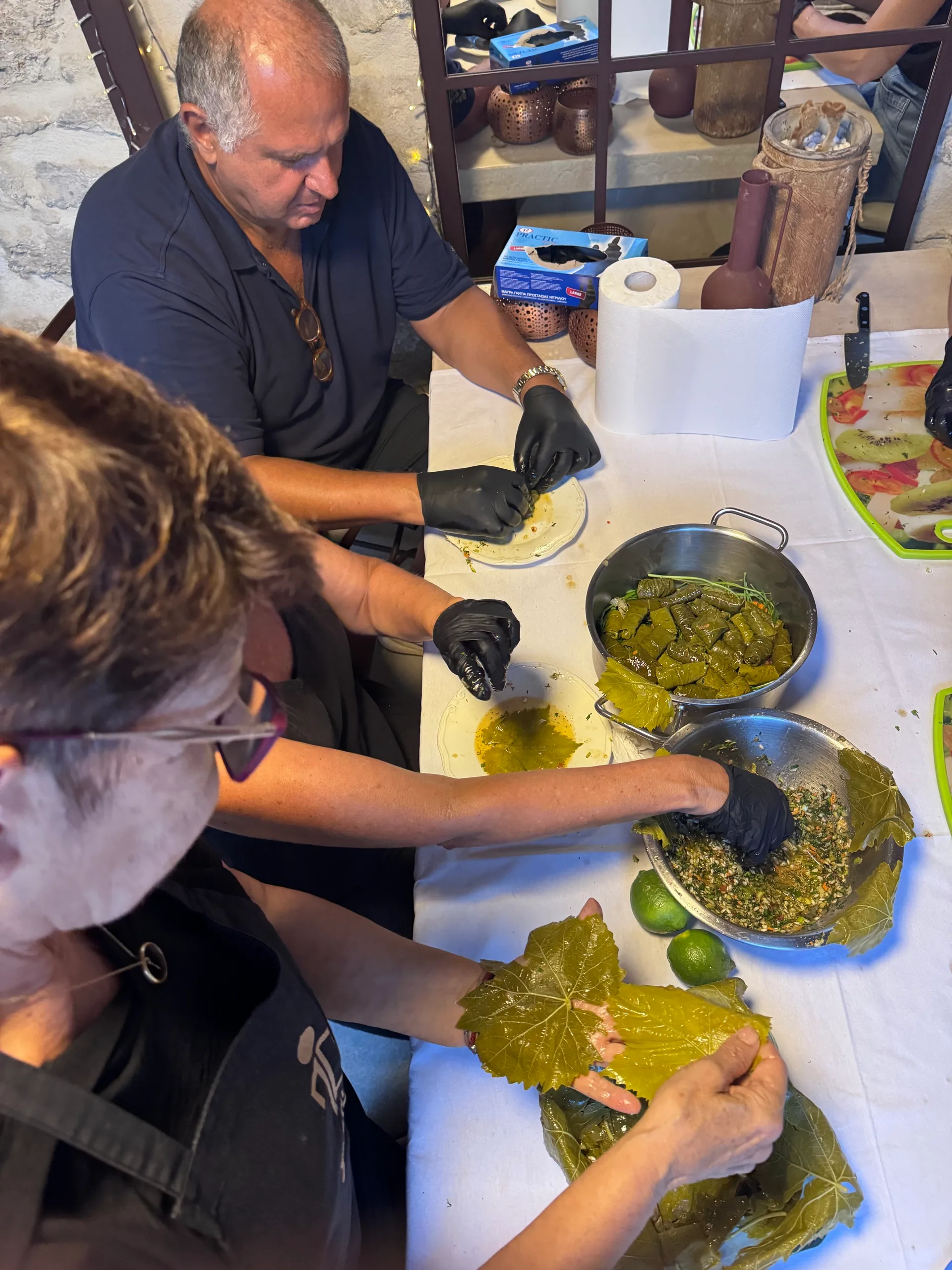 People filling and rolling grape leaves around a table with a pot of folded dolmathes