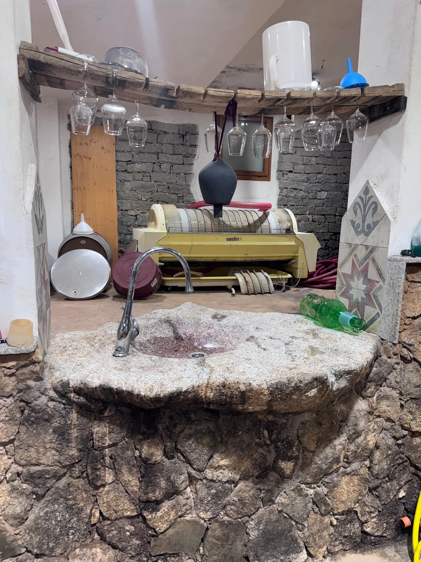 A rough-hewn granite sink with wine press and cellar tools behind it